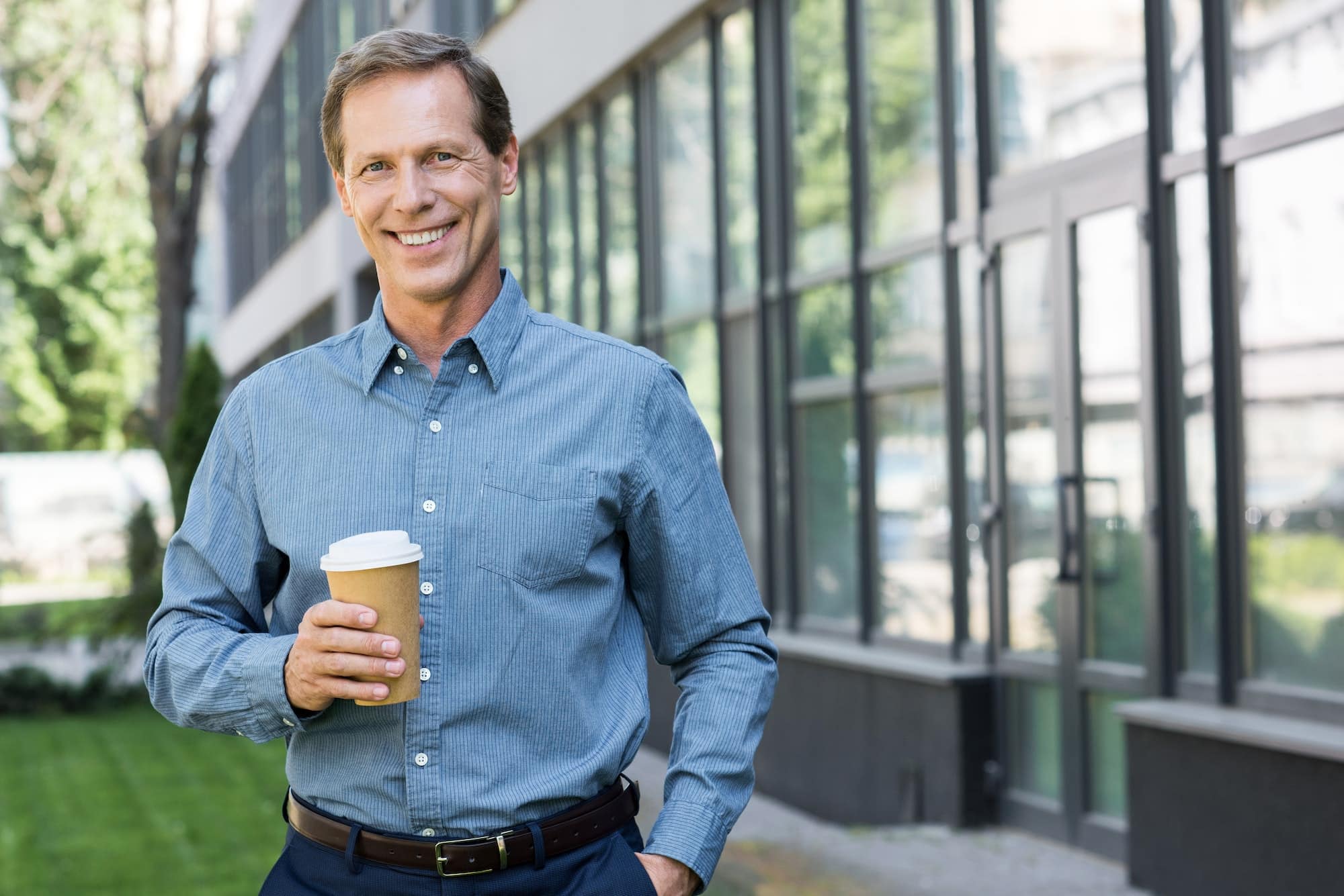 smiling businessman posing with disposable cup of coffee near office building